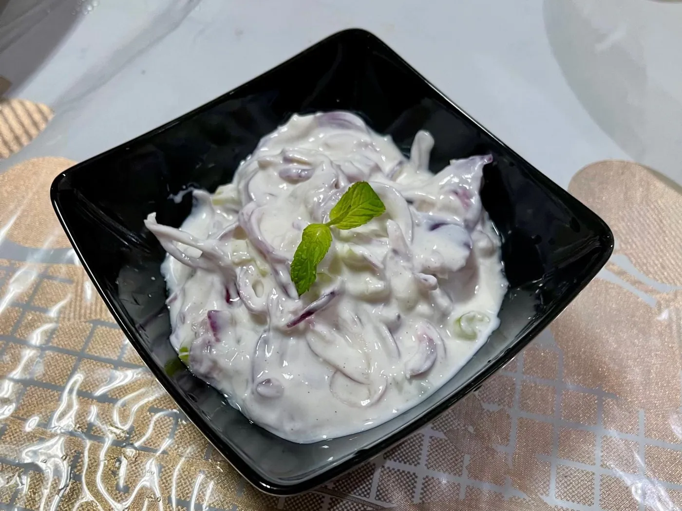 Creamy yogurt salad with thinly sliced red onion in a square black bowl, garnished with a sprig of mint