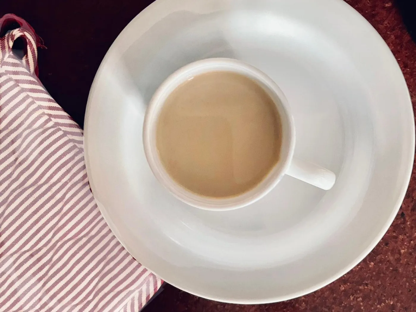 A white cup of creamy coffee milk tea on a white saucer beside a red-and-white striped napkin