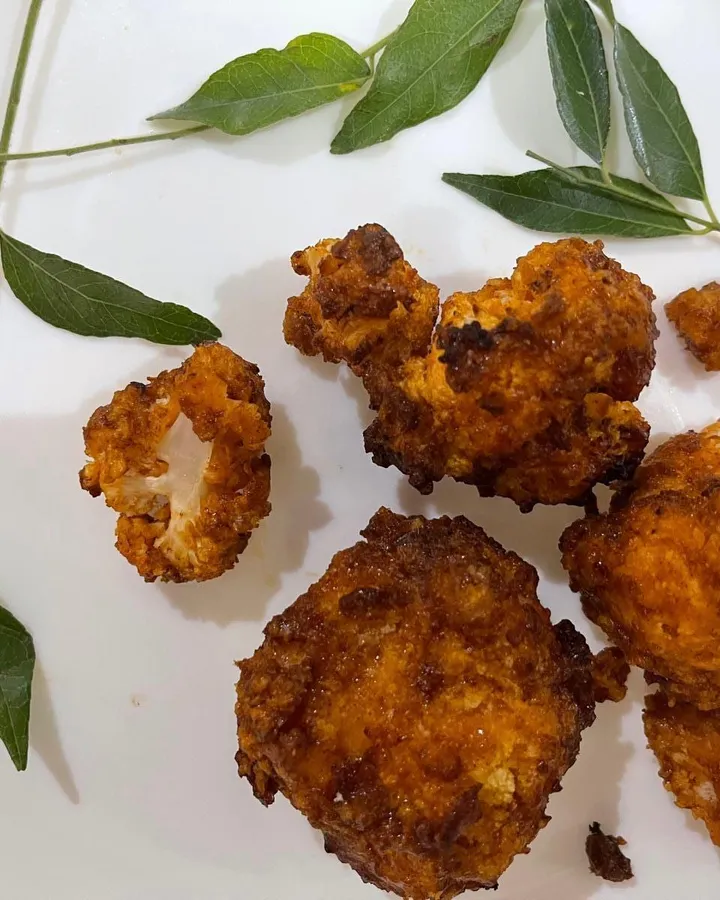 Crispy golden air-fried buffalo cauliflower bites on a white plate with sprigs of curry leaves