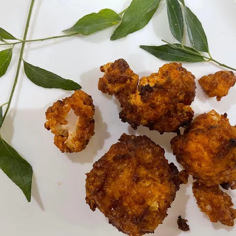 Crispy golden air-fried buffalo cauliflower bites on a white plate with sprigs of curry leaves