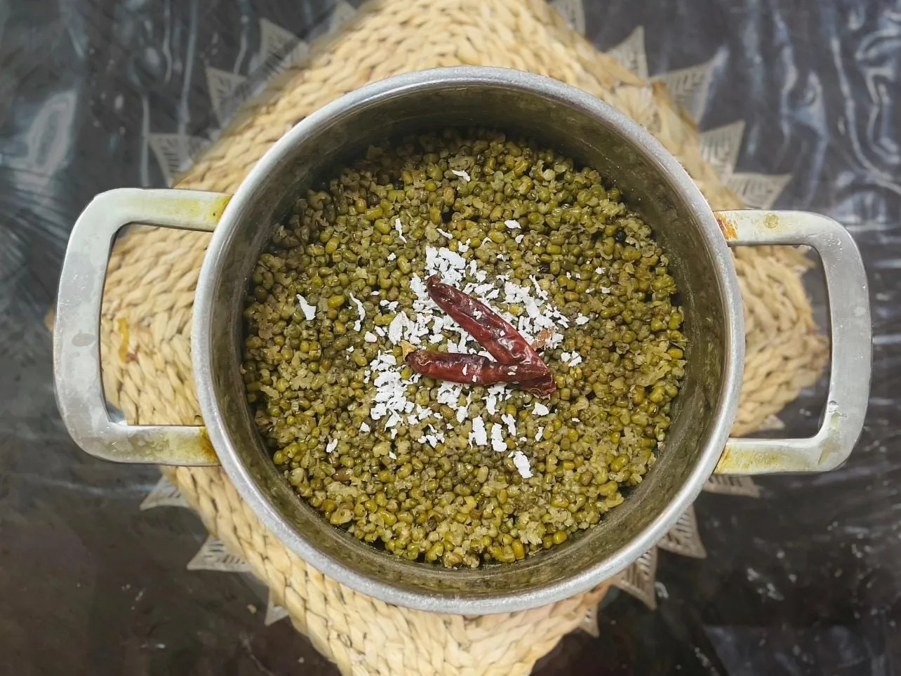 A pot of boiled green gram topped with scraped coconut and dried red chilies, on a woven mat