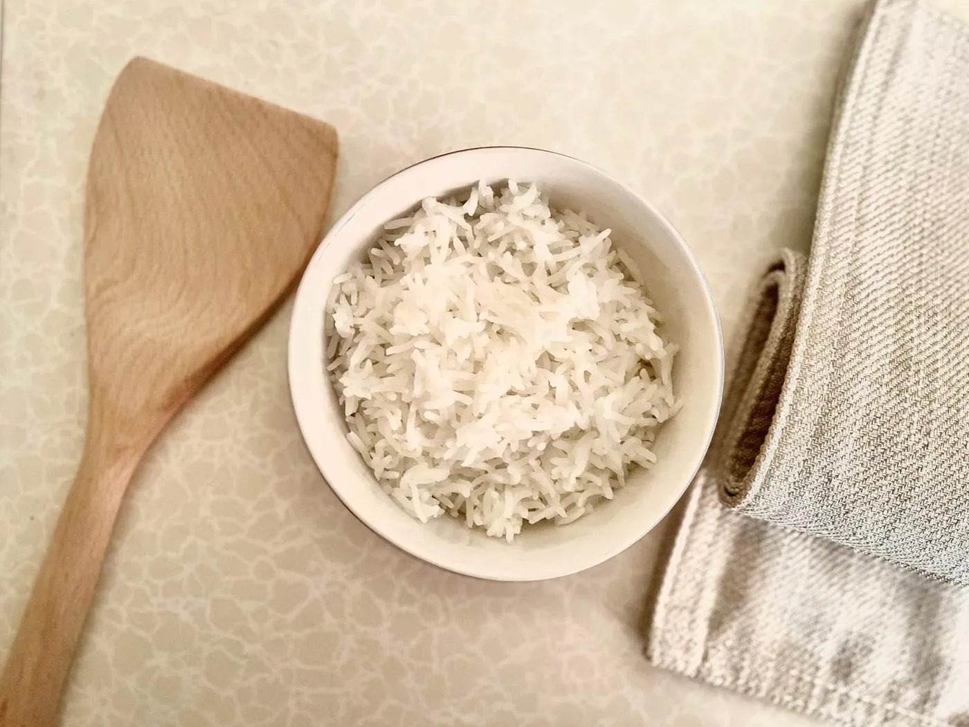 A white bowl of fluffy cooked white rice next to a wooden spatula and a linen napkin
