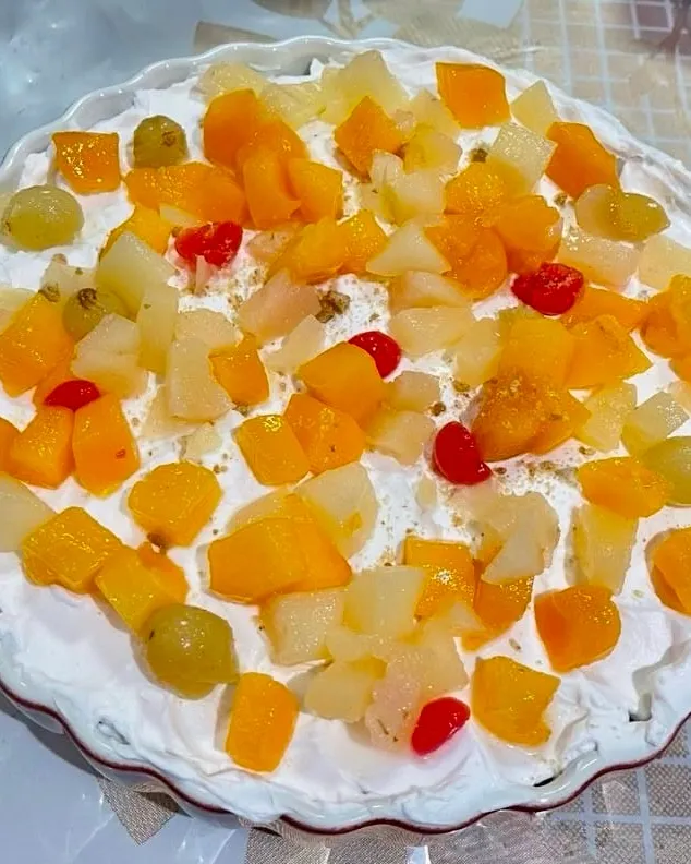 Fruit custard trifle in a white fluted dish, topped with whipped cream and colorful mixed fruit